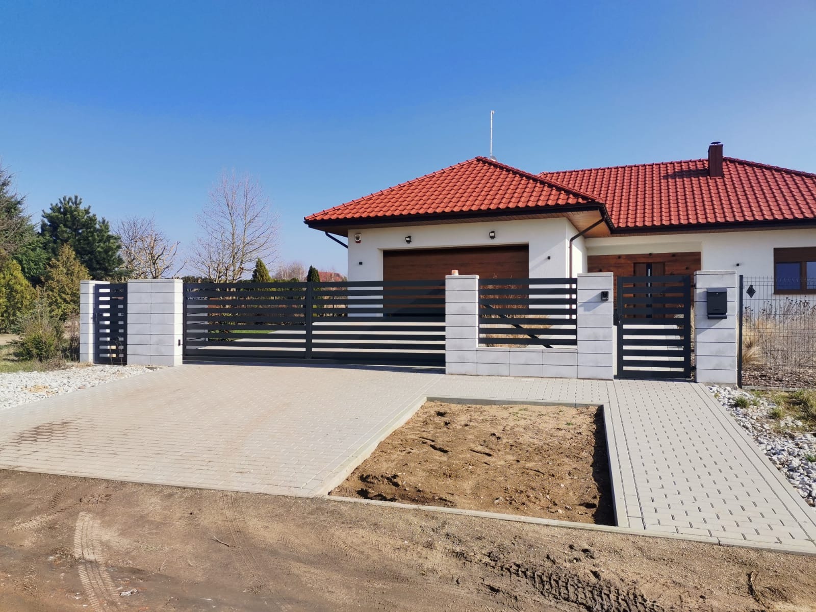 Nowoczesna, pozioma, czarna brama wjazdowa z przesuwnym skrzydłem i furtką, osadzona na murowanych, szarych słupkach z betonu architektonicznego, na tle domu z czerwoną dachówką i podjazdem...