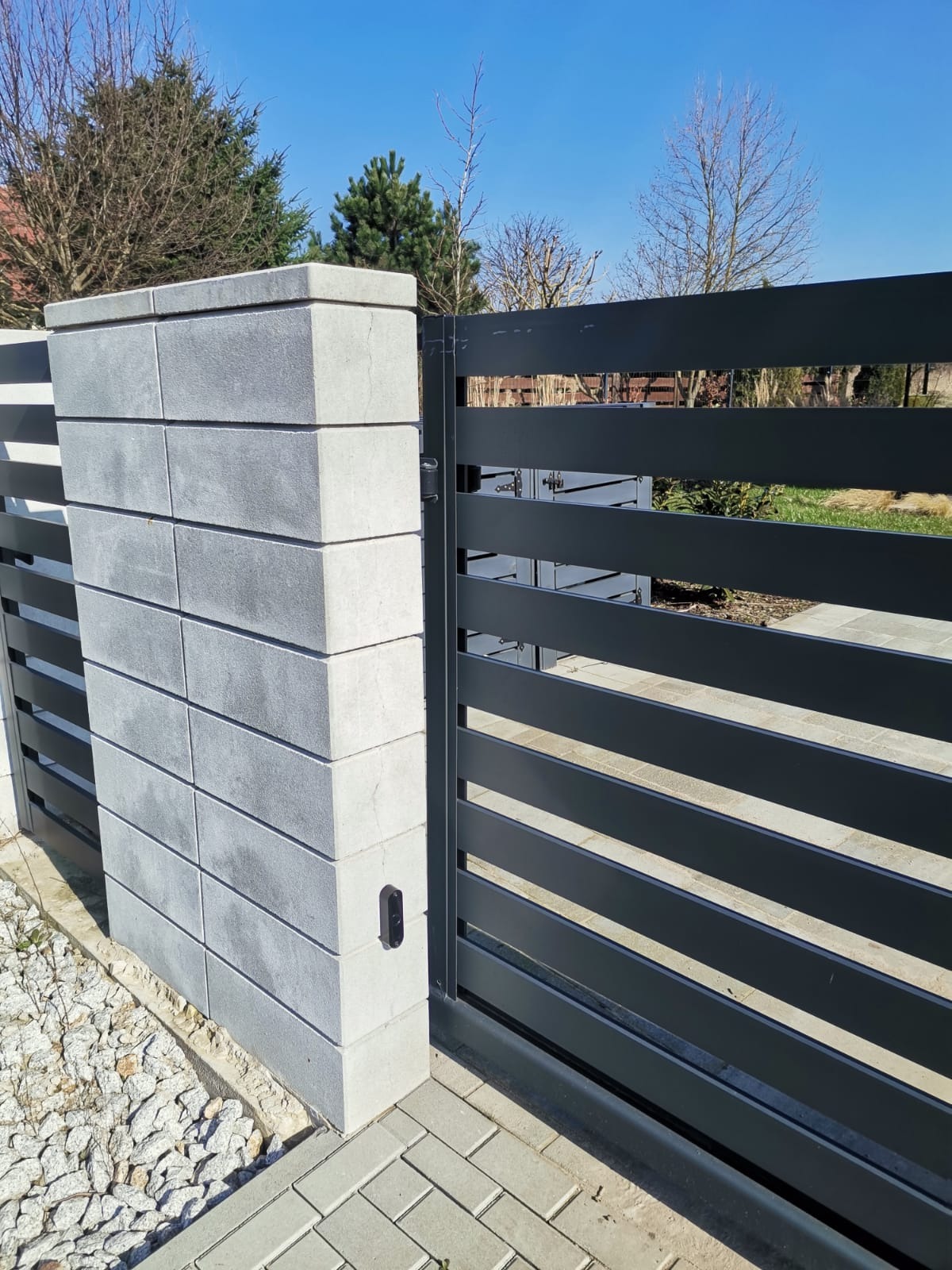 Nowoczesna, pozioma brama w kolorze antracytowym z betonowym słupkiem z bloczków, widoczny czytnik zbliżeniowy, kostka brukowa i biały żwir u podstawy.
