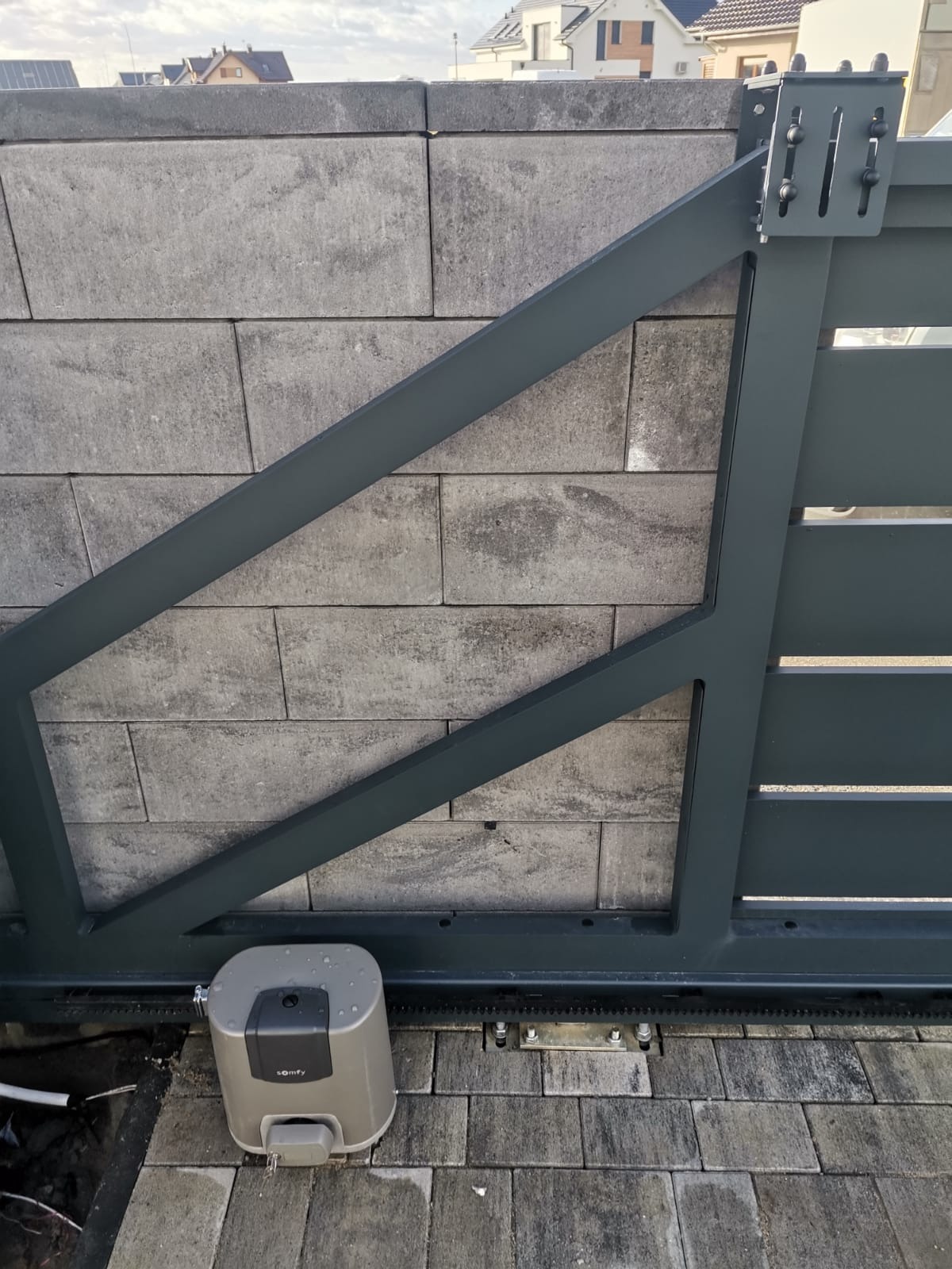 Szara brama przesuwna z napędem Somfy, zamontowana na tle betonowego muru z bloków, widoczna z poziomu gruntu.