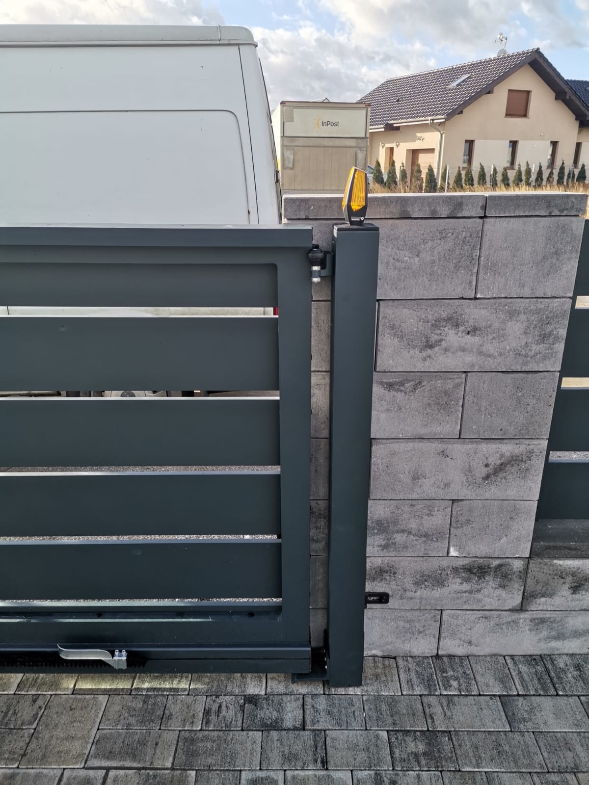 Szara brama przesuwna z poziomymi panelami, zamontowana na słupku z szarej kostki betonowej, z widoczną lampą ostrzegawczą, z tyłu biały bus i paczkomat InPost.