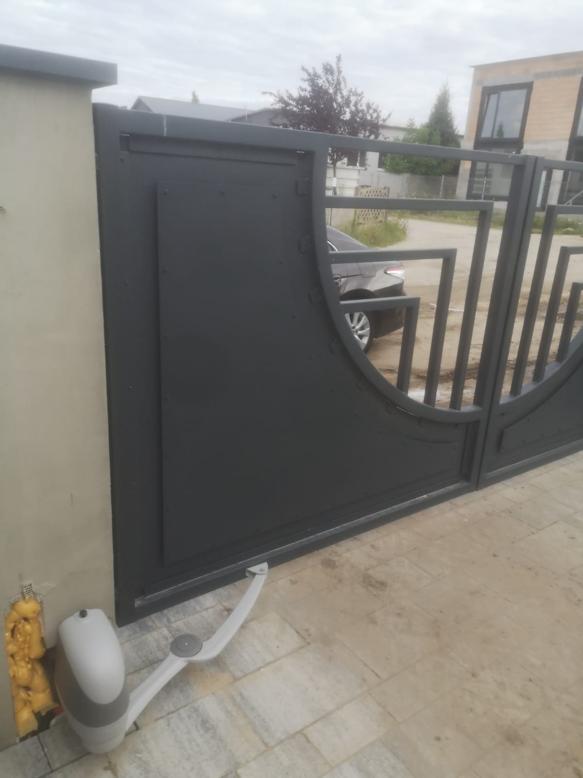 Szara brama wjazdowa z automatyką, widoczny siłownik przy słupku, na podjeździe kostka brukowa, w tle samochód osobowy i budynek.