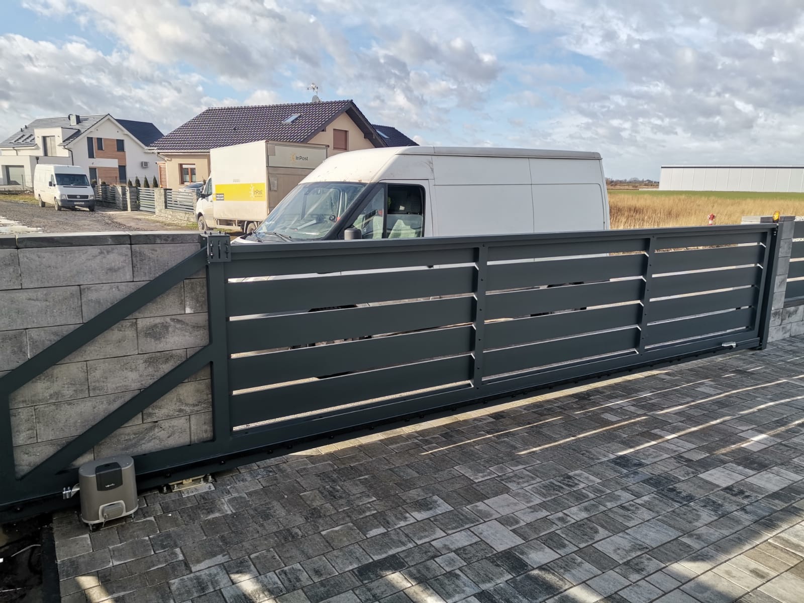 Nowoczesna, przesuwna brama wjazdowa z poziomymi panelami w kolorze antracytowym, zamontowana na murowanych słupkach z szarej kostki, z widocznym napędem automatycznym i dwoma samochodami...