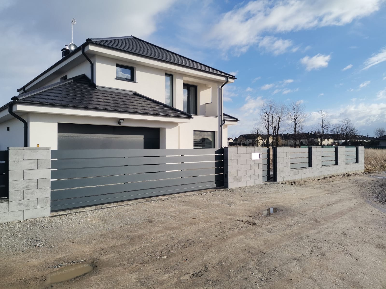 Nowoczesny dom z szarą bramą przesuwną i ogrodzeniem z betonowych bloczków, widoczne kałuże na gruncie.