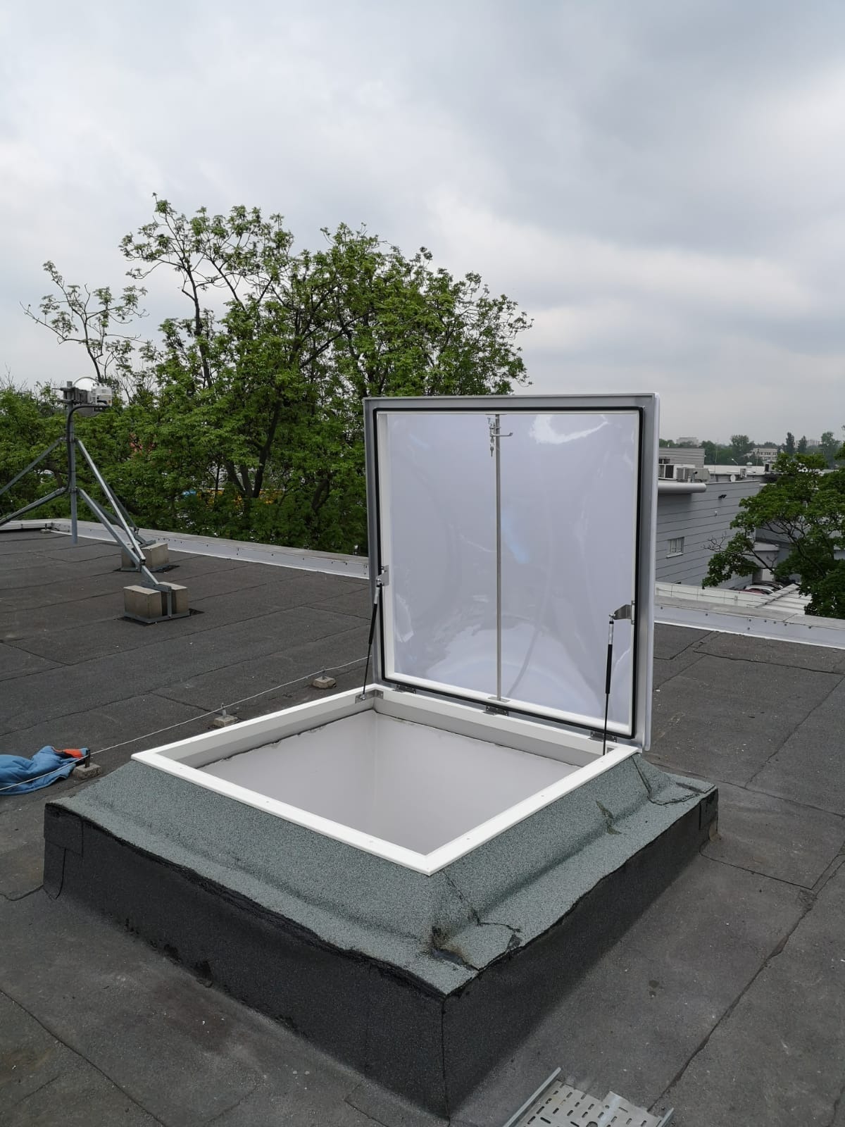 Otwarty świetlik dachowy z białą ramą na płaskim dachu pokrytym papą, widoczne mechanizmy podtrzymujące klapę, w tle drzewa i budynki miejskie pod pochmurnym niebem.