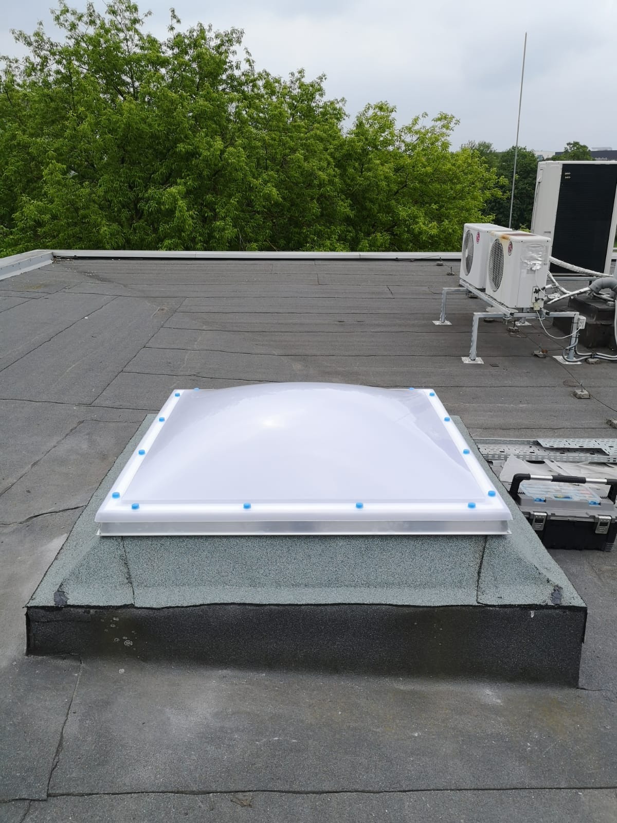 Kopuła doświetlająca zamontowana na płaskim dachu pokrytym papą, widoczna skrzynka narzędziowa i klimatyzatory w tle.