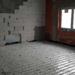 Rozłożone rury ogrzewania podłogowego na folii w surowym stanie, widoczne fragmenty ścian z pustaków i dwa okna z białymi ramami.