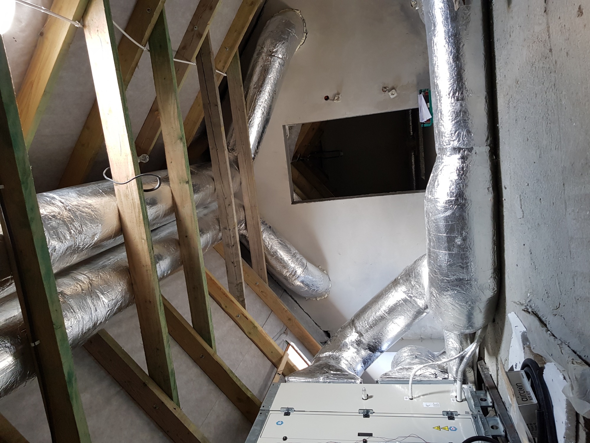 Instalacja wentylacyjna z aluminiowymi przewodami w surowym wnętrzu poddasza, widoczne drewniane elementy konstrukcyjne dachu i biała centrala wentylacyjna z przewodami.