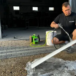 Mężczyzna w czarnej koszulce z napisem 'R-KLIMA' czyści białą listwę za pomocą myjki ciśnieniowej na zewnątrz budynku, widoczny zielony agregat i wiadro z wodą.