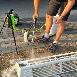 R-KLIMA Rafał Maćków - Czyszczenie obudowy klimatyzatora wysokociśnieniową myjką na zewnątrz, widoczny pracownik w sportowych butach New Balance, a obok na trójnogu stoi zielona myjka ciśnieniowa z wyświetlaczem.