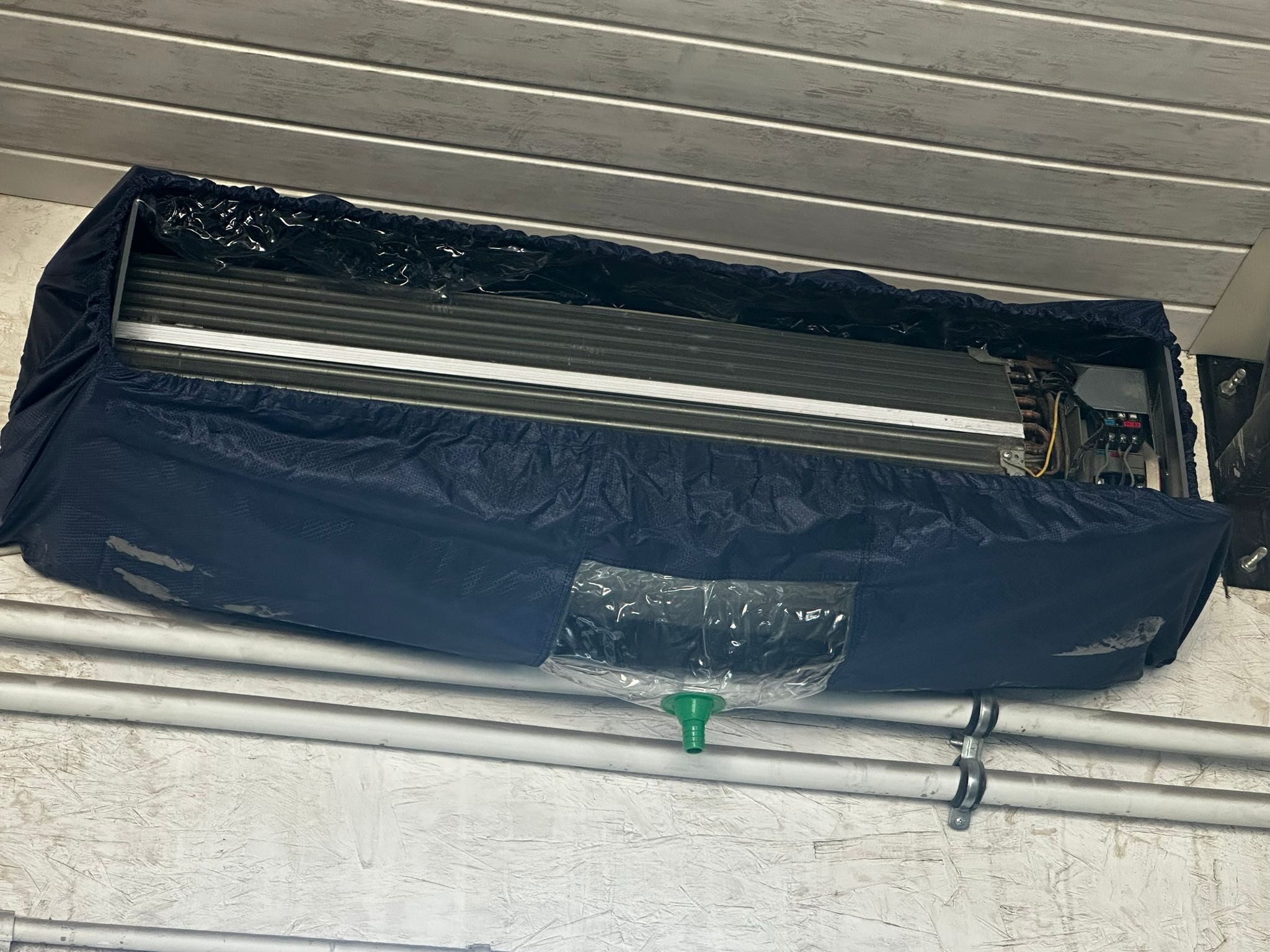 Klimatyzator domowy zabezpieczony niebieską osłoną z przezroczystym odpływem skroplin, widoczne rury instalacyjne na tle ściany z boazerią.