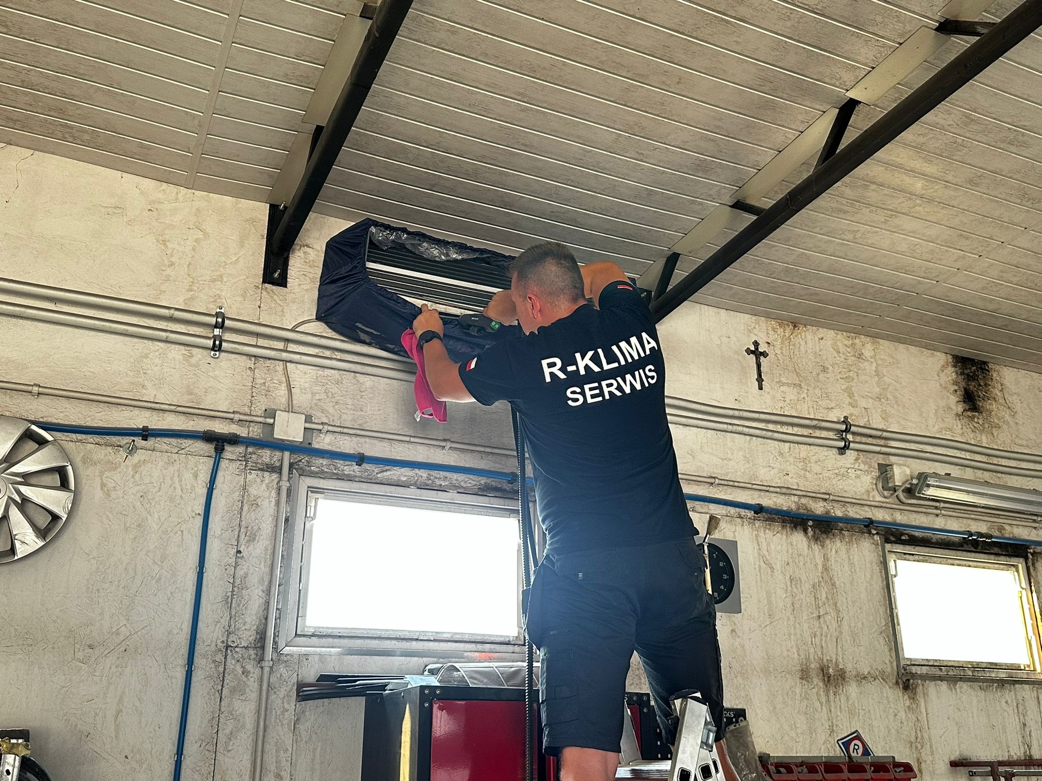 Mężczyzna w czarnej koszulce z napisem 'R-KLIMA SERWIS' czyści klimatyzator ścienny w pomieszczeniu warsztatowym, stojąc na aluminiowej drabinie.
