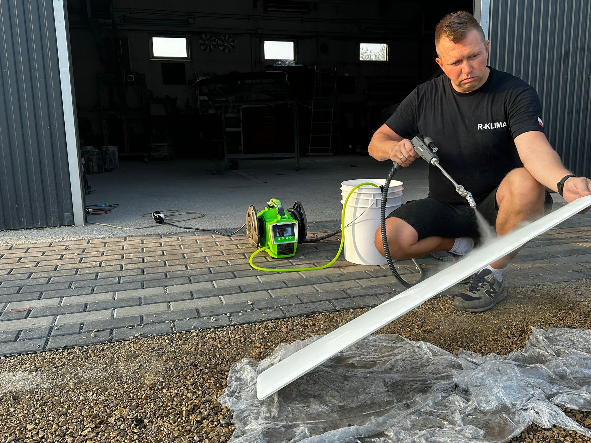 Mężczyzna w czarnej koszulce z napisem 'R-KLIMA' czyści białą listwę za pomocą myjki ciśnieniowej na zewnątrz budynku, widoczny zielony agregat i wiadro z wodą.