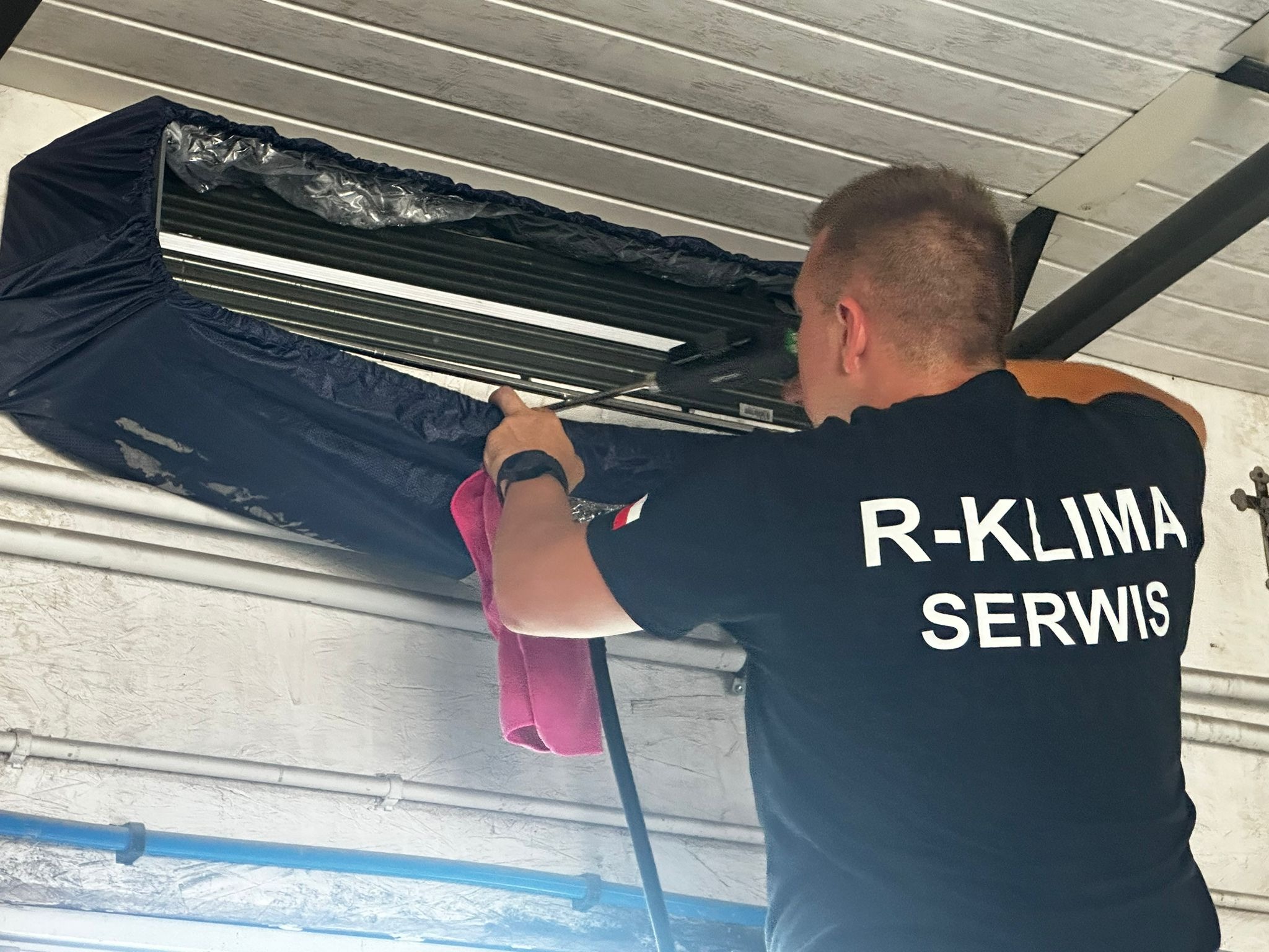 Mężczyzna w czarnej koszulce z napisem 'R-KLIMA SERWIS' czyści klimatyzator za pomocą szczotki i ściereczki, zabezpieczony pokrowcem z folią.