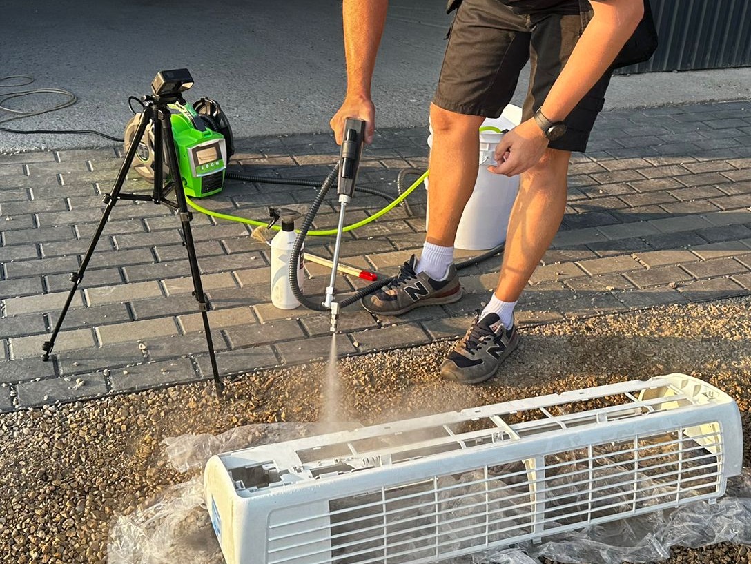 Czyszczenie obudowy klimatyzatora wysokociśnieniową myjką na zewnątrz, widoczny pracownik w sportowych butach New Balance, a obok na trójnogu stoi zielona myjka ciśnieniowa z wyświetlaczem.