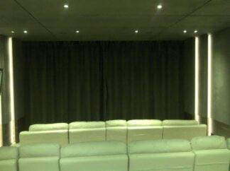 Kino domowe: rząd kremowych, skórzanych foteli przed zasłoniętym ekranem. Minimalistyczne oświetlenie po bokach, surowy beton na ścianach i suficie.