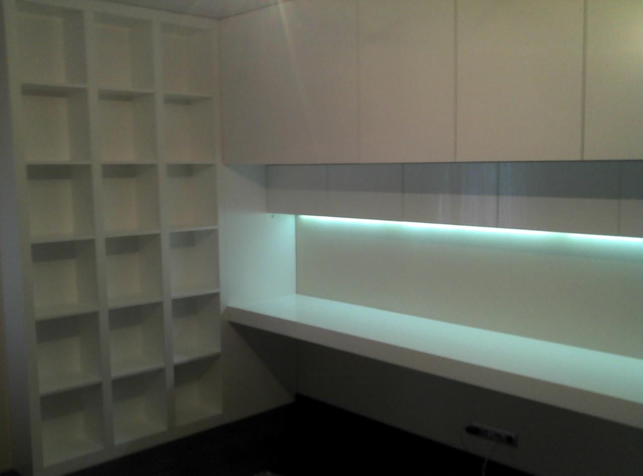 Biała zabudowa meblowa z podświetleniem LED: regał z kwadratowymi półkami i biurko z szafkami wiszącymi. Minimalistyczny design, nowoczesne oświetlenie.