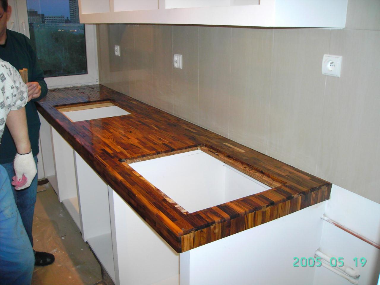 Montaż blatu z egzotycznego drewna w kuchni. Widoczne wycięcia na zlew i płytę grzewczą. Białe szafki i płytki ścienne. Praca w toku, dwie osoby w tle.