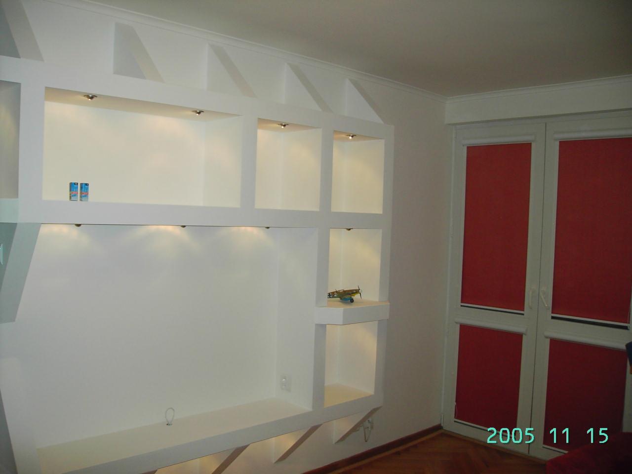 Biała zabudowa ścienna z półkami i oświetleniem halogenowym, widok na okno z czerwoną roletą, parkiet na podłodze. Minimalistyczny design, nowoczesne wykończenie.