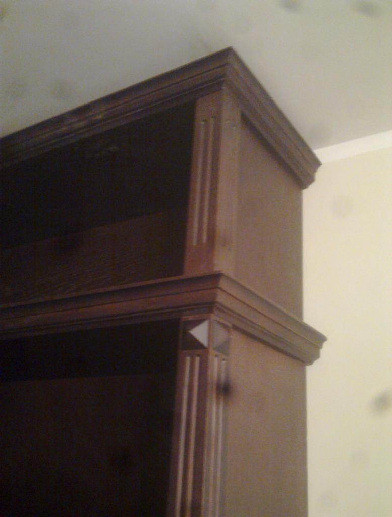 Fragment brązowej, drewnianej szafy z półkami, widoczny pod kątem od dołu, z dekoracyjnymi listwami i geometrycznym zdobieniem na rogu.