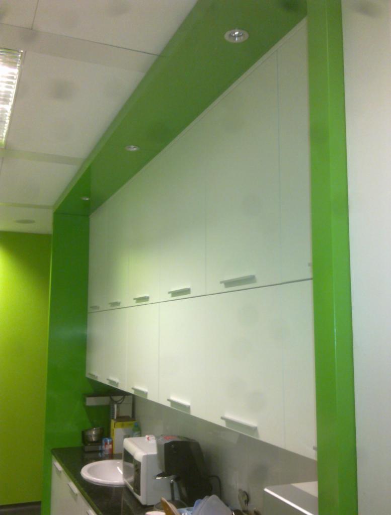 Białe szafki wiszące z poziomymi uchwytami w biurowej kuchni, z zielonymi akcentami kolorystycznymi na ścianach i obudowie. Widoczne wyposażenie AGD.