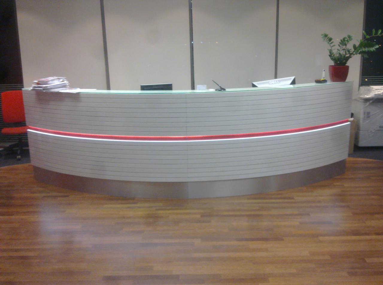 Biała, zaokrąglona recepcja z czerwoną listwą LED, drewniana podłoga. W tle ściana z paneli i roślina doniczkowa. Nowoczesny design przestrzeni biurowej.