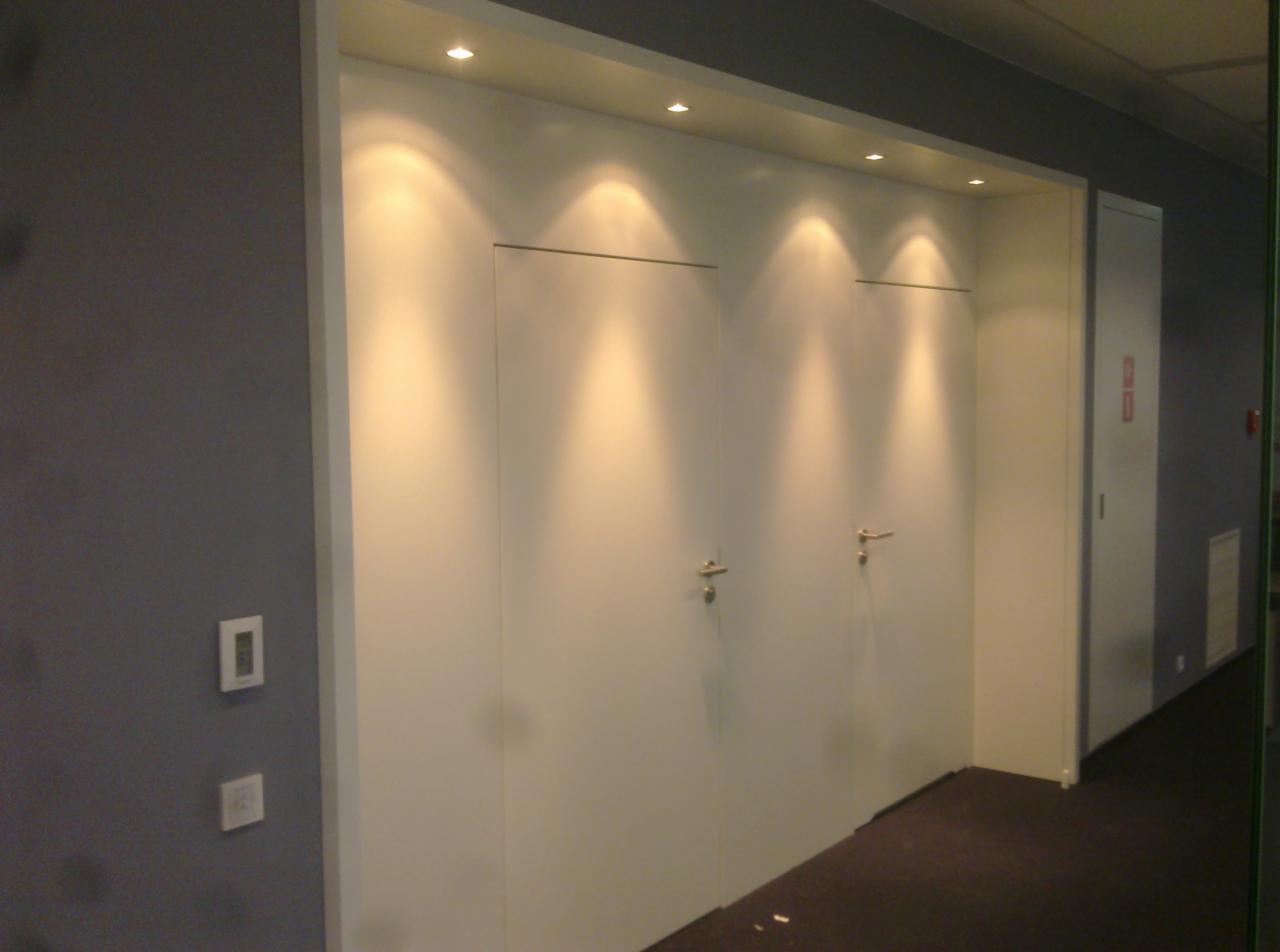 Dwie białe, ukryte drzwi z minimalistycznymi klamkami w długim korytarzu biurowym oświetlonym punktowymi lampami sufitowymi. Szara ściana z włącznikami.