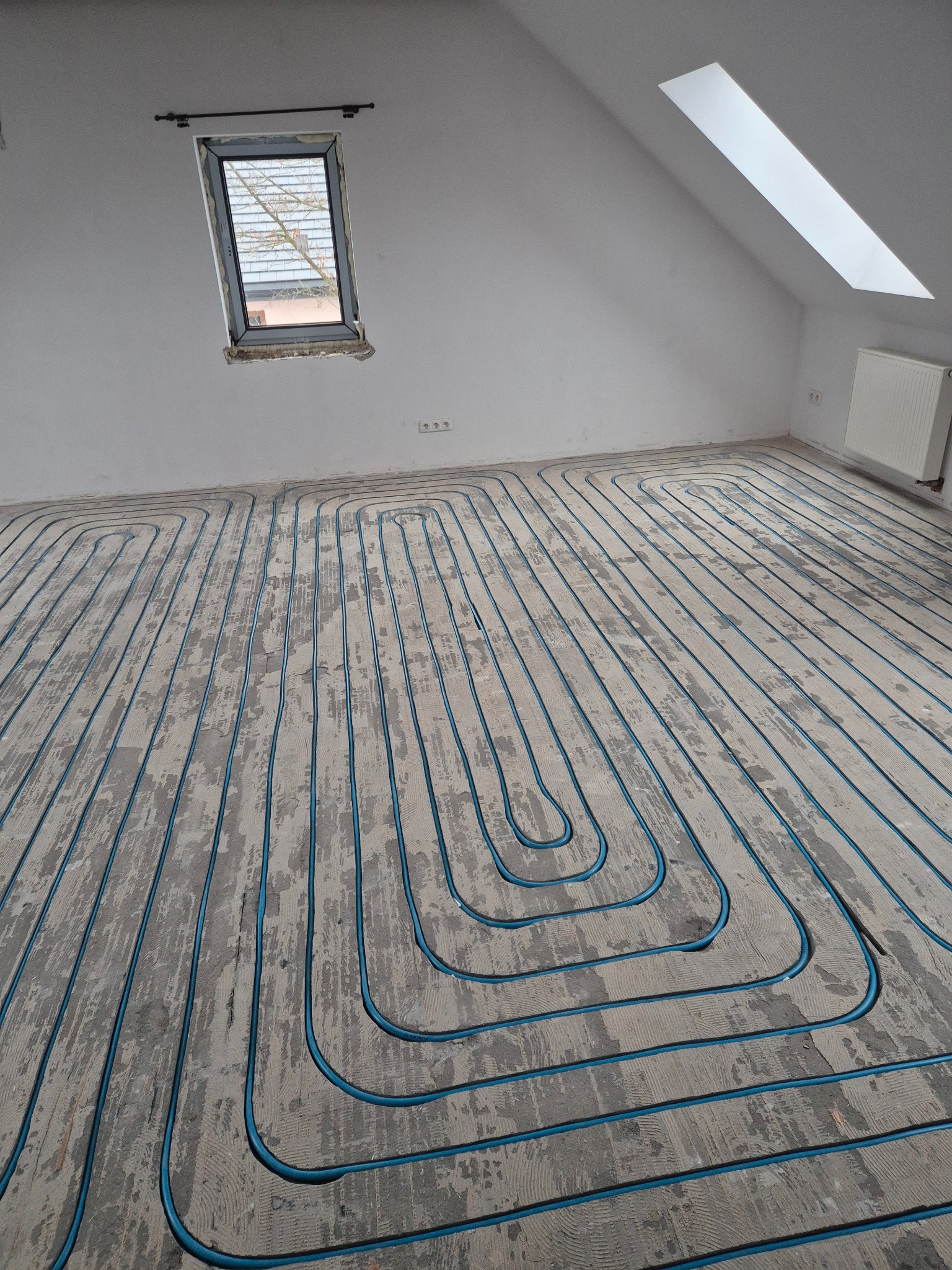 Niebieskie rury ogrzewania podłogowego ułożone w jodełkę na szarej, surowej posadzce w jasnym pokoju na poddaszu z oknami dachowymi i grzejnikiem.