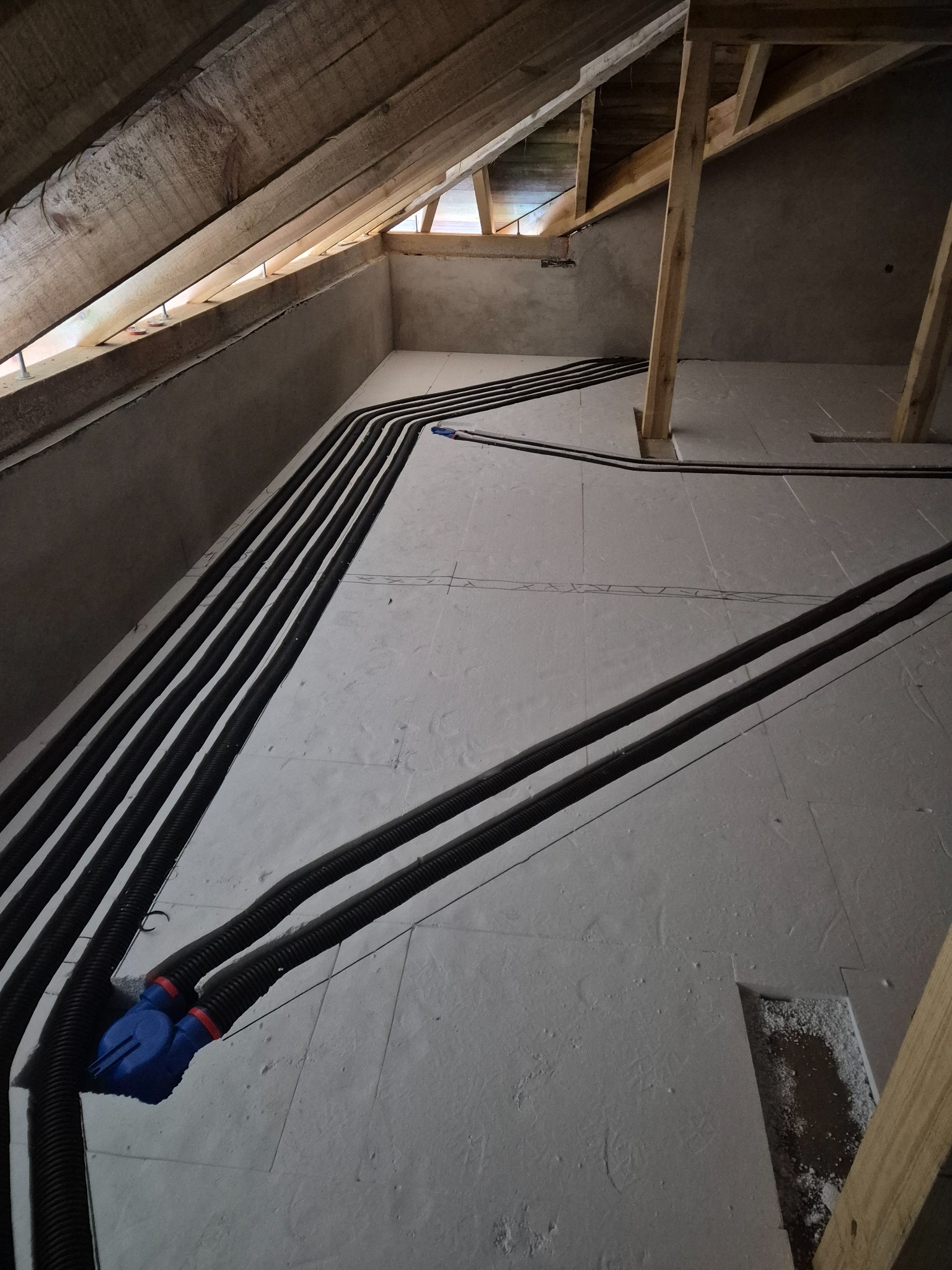 Czarny, karbowany system rur ułożony równolegle na szarej powierzchni poddasza, z widoczną drewnianą konstrukcją dachu i ścian. Fragment instalacji z niebieskim elementem.