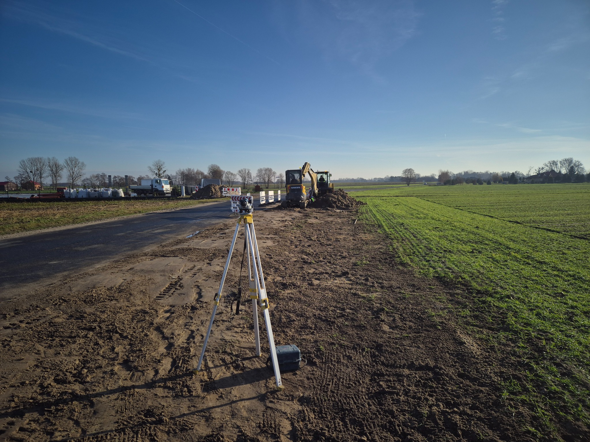Żółta koparka Volvo na placu budowy, obok niwelator optyczny. W tle droga, drzewa i błękitne niebo. Prace ziemne w toku, przygotowanie terenu pod inwestycję.