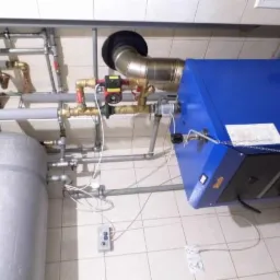 Niebieski piec centralnego ogrzewania z szarym zbiornikiem akumulacyjnym, rurami miedzianymi i stalowymi, oraz białą płytką na ścianie i podłodze.