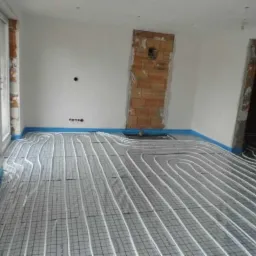 Wnętrze w trakcie instalacji ogrzewania podłogowego: białe rurki ułożone na szarej macie z niebieską taśmą brzegową, ściany w surowym stanie, widoczne fragmenty cegieł.