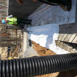 Instalacja rury drenażowej w wykopie zabezpieczonym geowłókniną, widoczny pracownik w kasku i kamizelce odblaskowej oraz fragment zbrojenia konstrukcji betonowej.