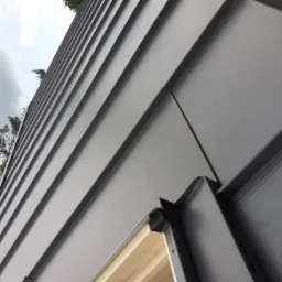 Szare panele blaszane na ścianie budynku, widoczne połączenia i fragment drewnianej konstrukcji pod spodem, ujęcie z dołu.