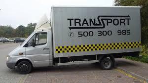 Srebrny samochód dostawczy z nadrukowanym napisem 'TRANSPORT' i numerem telefonu, zaparkowany na ulicy. Na boku pojazdu widoczny pas w czarno-żółtą szachownicę.