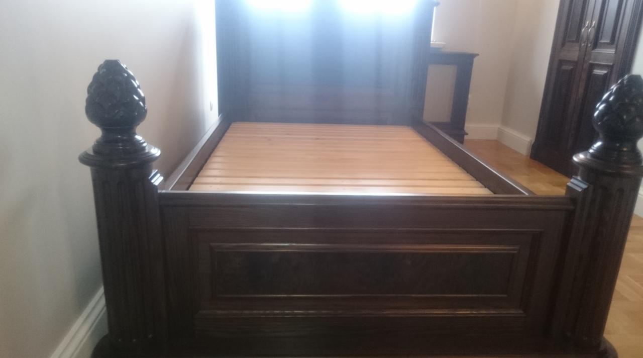 Ciemnobrązowe, drewniane łóżko z dekoracyjnymi zwieńczeniami w kształcie szyszki, widoczne na tle jasnej ściany i podłogi, z szafą w tle.