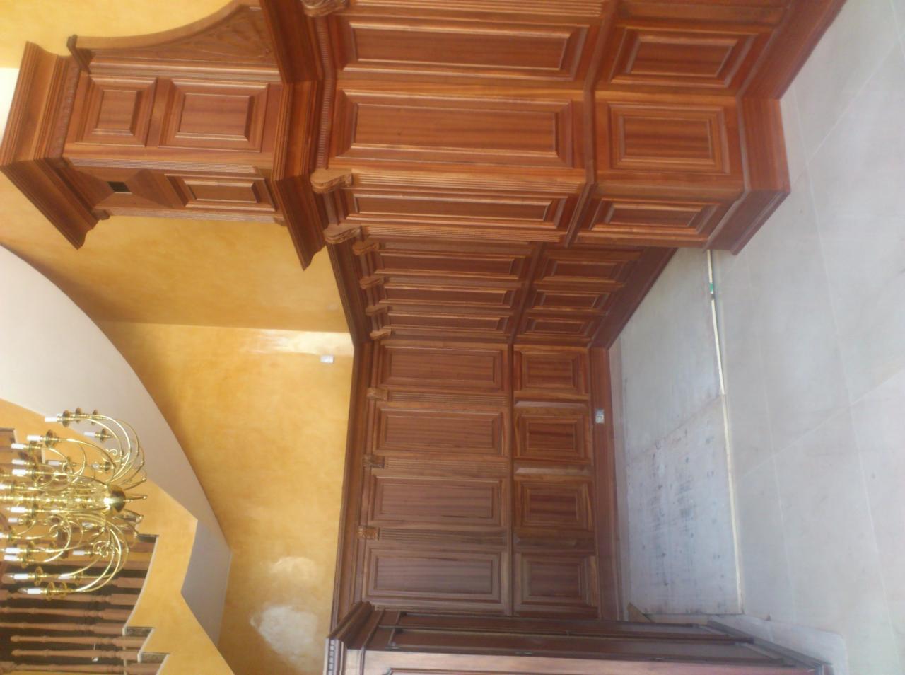 Drewniana zabudowa sufitu z kasetonami i ozdobnymi listwami, widok z dołu, oświetlona złotym światłem żyrandola, w tle ściana w ciepłym odcieniu żółci.