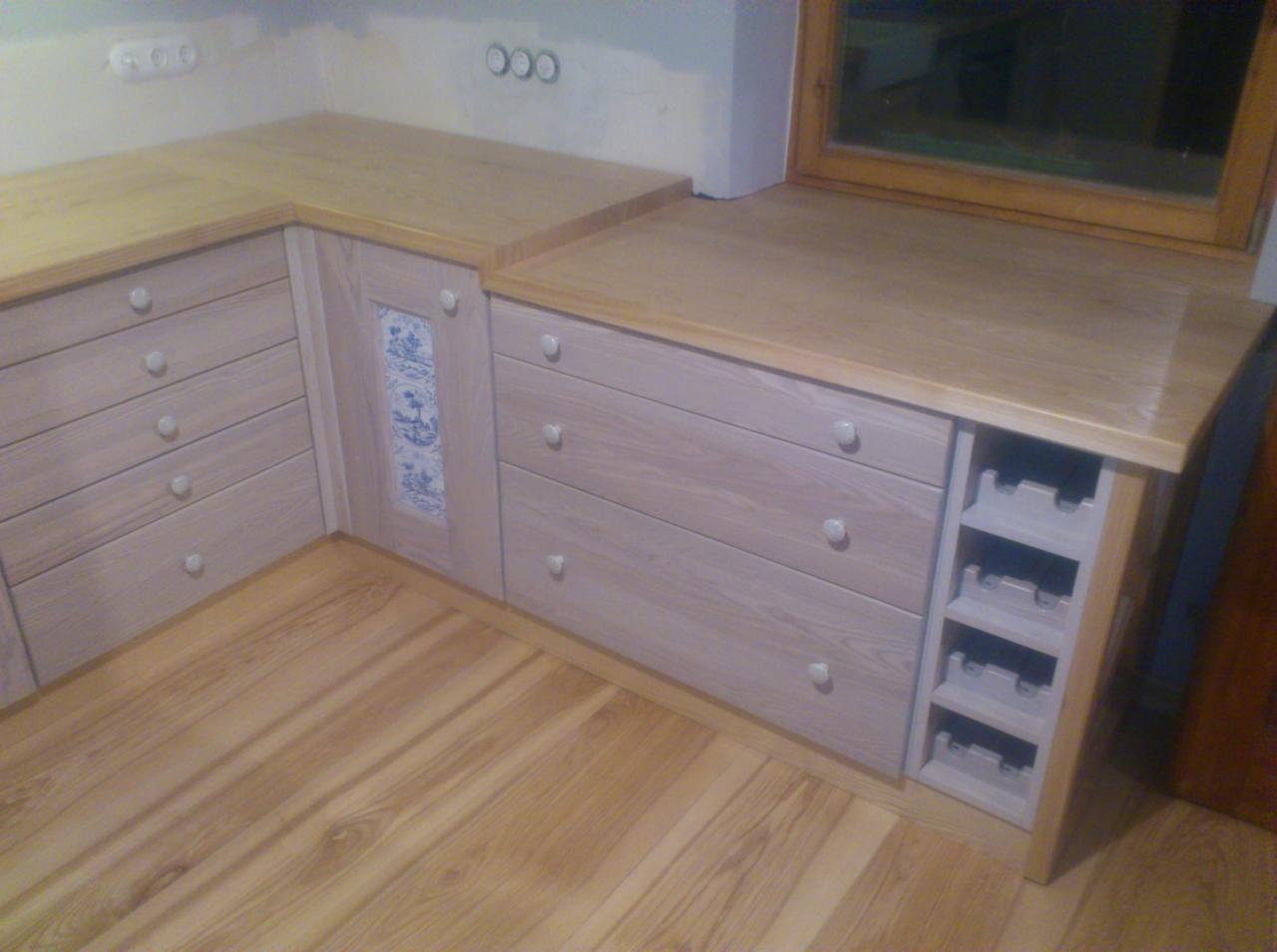 Drewniana zabudowa meblowa z szufladami, jasne drewno, białe gałki, dekoracyjny panel z motywem kwiatowym, wnęka na butelki, parkiet na podłodze.