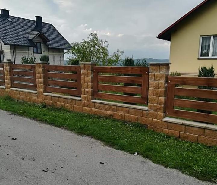 Nowoczesne ogrodzenie z drewnianymi przęsłami i elementami z imitacji kamienia, widok z poziomu ulicy, dwa domy w tle.