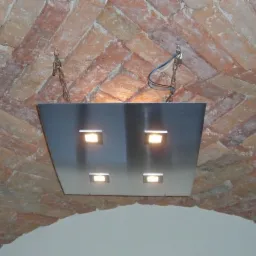 Metalowy panel oświetleniowy z czterema punktami świetlnymi zawieszony na łańcuchach pod ceglanym sklepieniem łukowym.