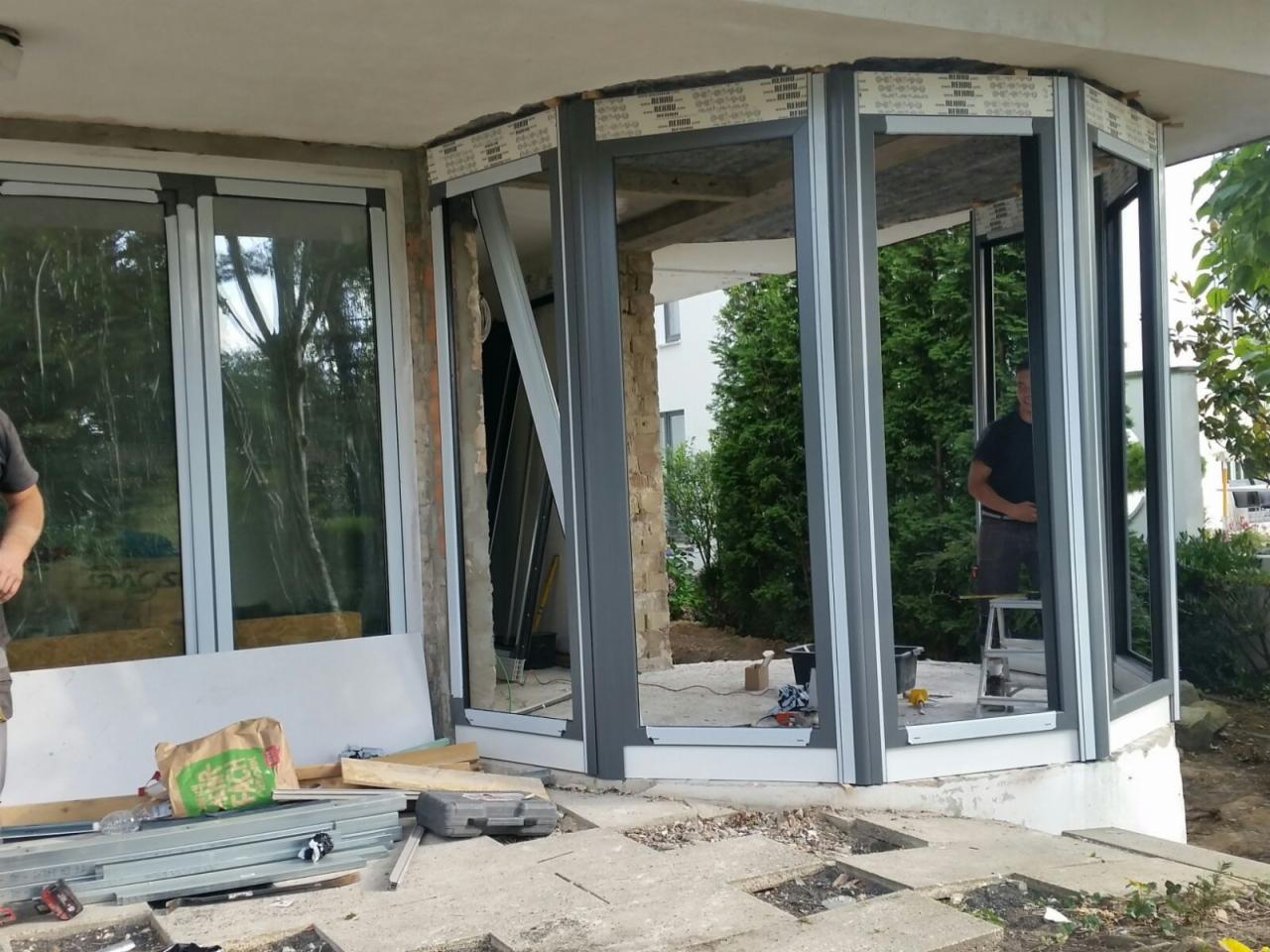 Montaż szarej, wielokątnej konstrukcji okiennej PCV na betonowym fundamencie, widoczne narzędzia, niezabudowane otwory okienne i pracownik na drabinie w tle.