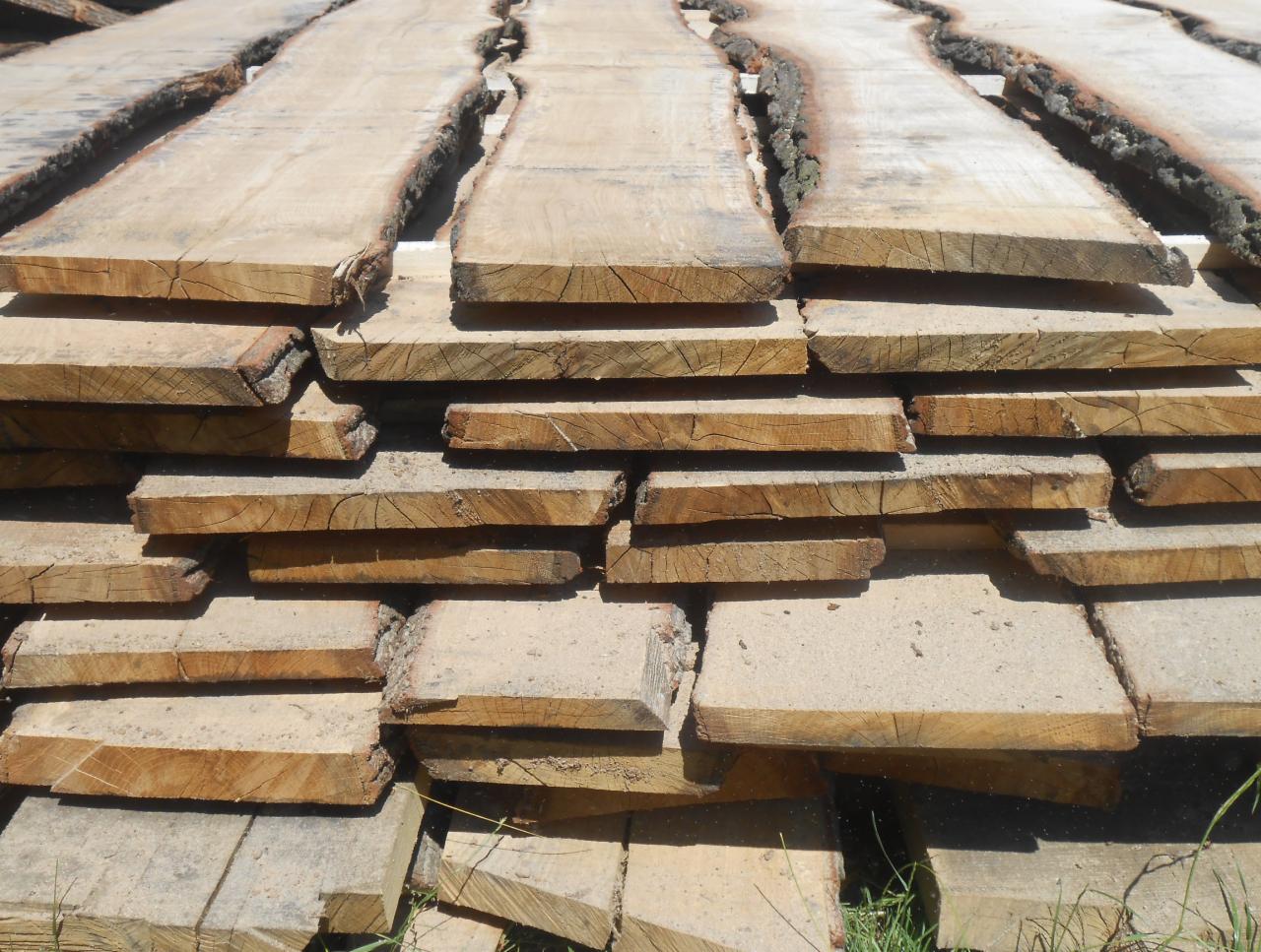 Ułożone deski tarcicy z korą na krawędziach, widoczne słoje drewna i struktura. Jasne, naturalne oświetlenie podkreśla detale drewna.