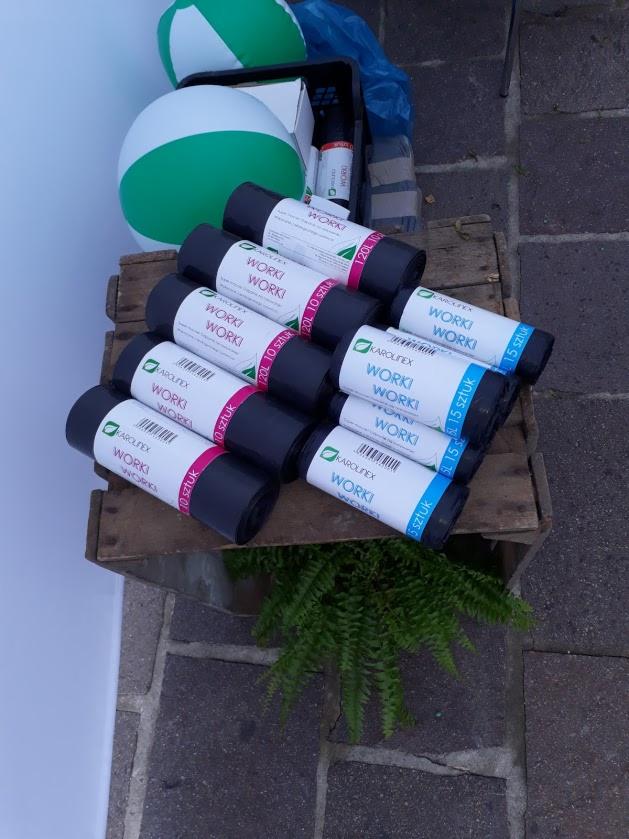 Zestaw rolek worków na śmieci Karolinex Worki 20L i 120L (10 sztuk) oraz 5L (15 sztuk) ułożonych na drewnianej skrzynce z zieloną rośliną ozdobną, w tle dmuchane piłki plażowe.