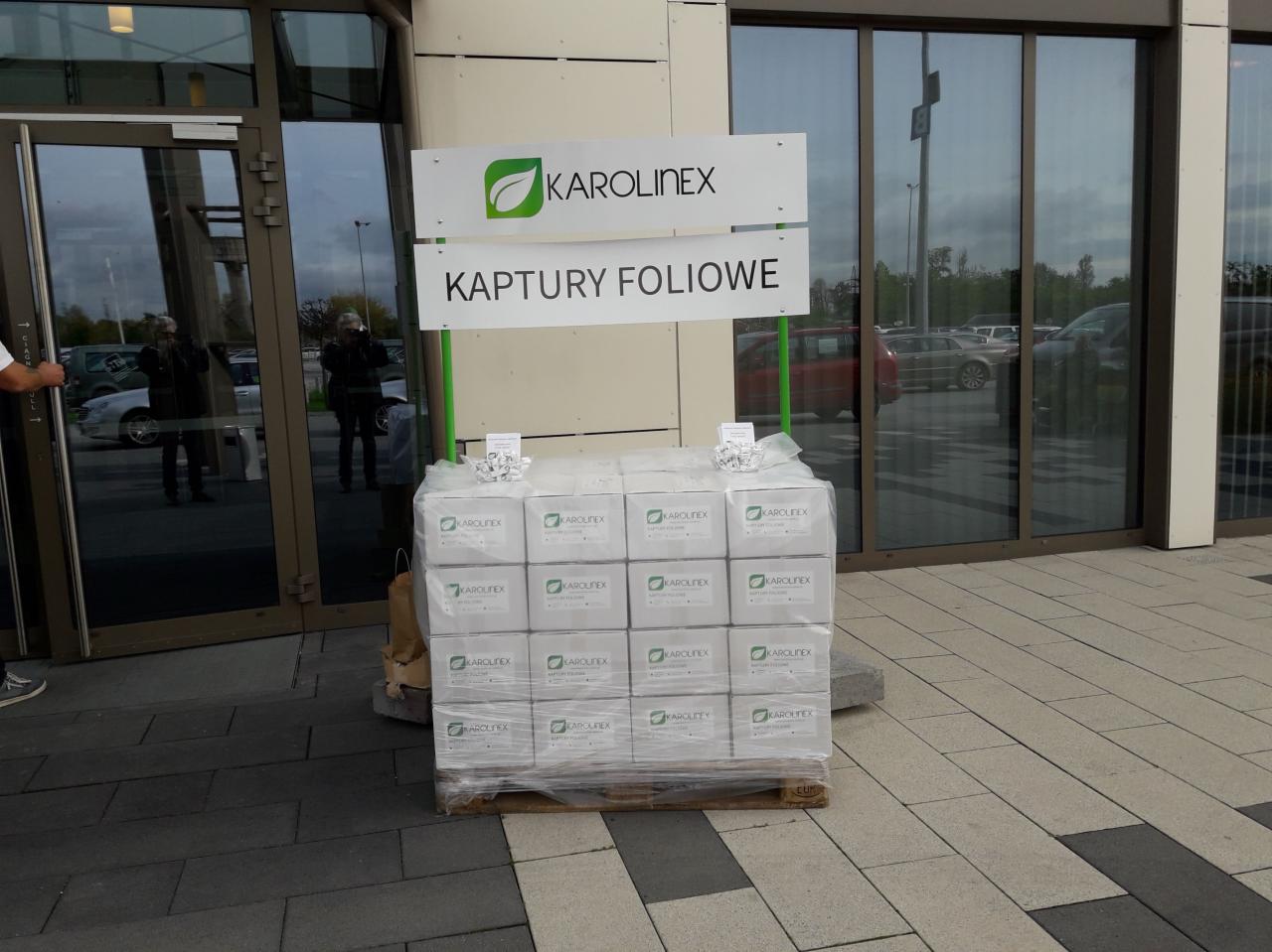 Paleta z zapakowanymi pudełkami z napisem KAROLINEX i KAPTURY FOLIOWE, ustawiona przed wejściem do budynku z przeszklonymi drzwiami.
