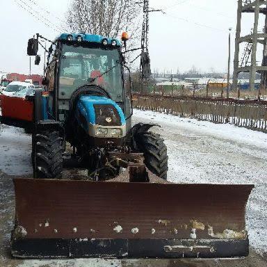 Niebieski traktor z pługiem do odśnieżania, stoi na ośnieżonej drodze, widoczne budynki w tle i słupy wysokiego napięcia.