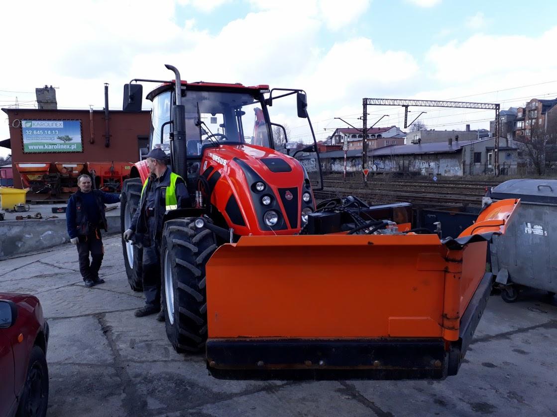 Czerwony traktor Ursus z pługiem śnieżnym, stojący na placu w pobliżu torów kolejowych, obok dwóch mężczyzn w roboczych ubraniach i żółtej kamizelce.