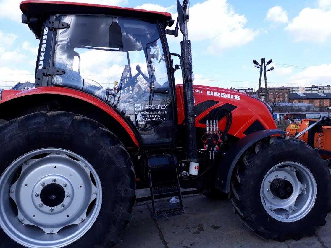 Czerwony ciągnik Ursus z widocznym logo firmy Karolinex na szybie kabiny, zaparkowany na zewnątrz w słoneczny dzień.