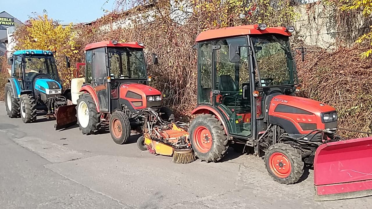 Trzy traktory komunalne (niebieski i dwa czerwone) z osprzętem do odśnieżania i czyszczenia ulic stoją zaparkowane na tle jesiennej roślinności.