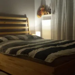 Drewniane łóżko z listwowym zagłówkiem w sypialni. Na ścianie wiszą zasłony i pluszowy miś. Lampka nocna na szafce obok łóżka tworzy przytulny nastrój.