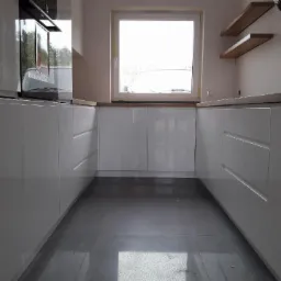 Nowoczesna, biała zabudowa kuchenna z połyskującymi frontami, drewnianym blatem i dwiema półkami na ścianie, widziana z niskiej perspektywy.