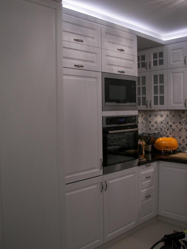 Biała zabudowa kuchenna z frezowanymi frontami, szafkami wiszącymi z przeszkleniami, piekarnikiem i mikrofalówką w zabudowie, oraz dekoracyjnym panelem nad blatem.